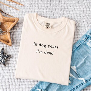 May include: A cream-coloured t-shirt with the text "in dog years I'm dead" in black. The shirt is folded and placed on a textured surface next to a pair of light blue denim jeans with distressed details.