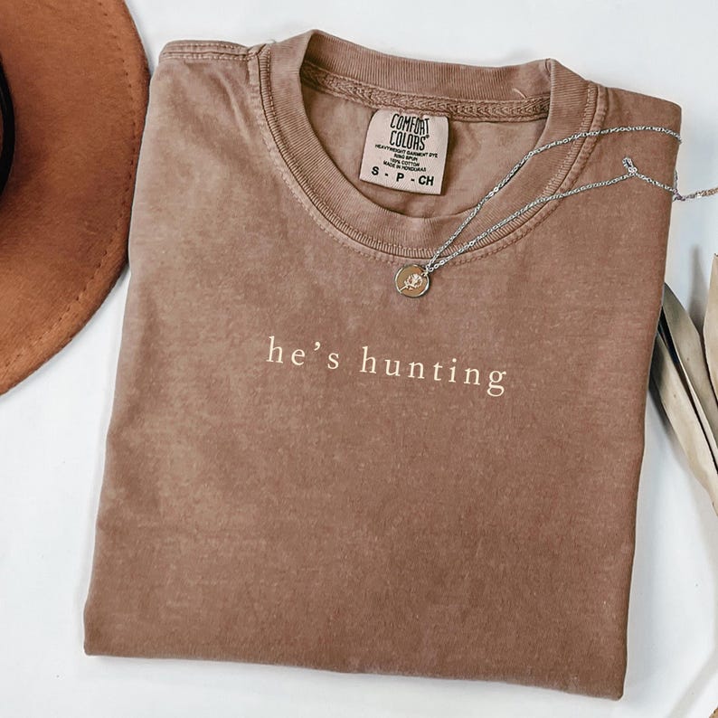 May include: A brown Comfort Colors t-shirt with the text "he's hunting" in a simple font. A silver necklace with a small pendant rests on the shirt. The shirt is folded and has a tag.