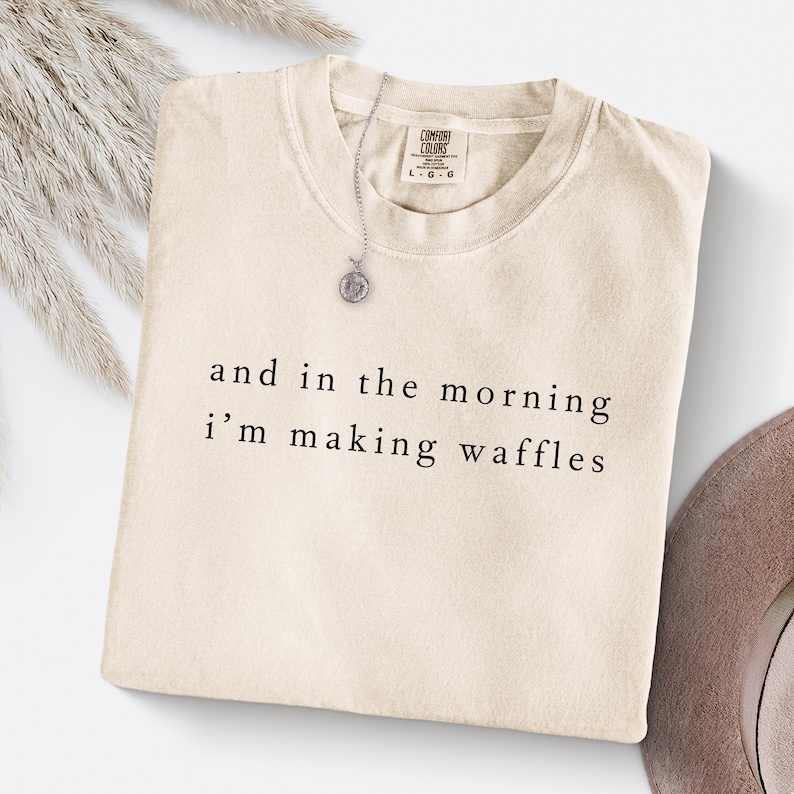 K&ouml;nnte beinhalten: Cremefarbenes Comfort Colors T-Shirt mit dem schwarzen Aufdruck "and in the morning i'm making waffles". Eine silberne Halskette mit rundem Anh&auml;nger liegt auf dem gefalteten Shirt.