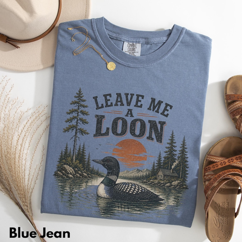 Pu&ograve; includere: Una t-shirt color blu jeans con una grafica raffigurante un strolaga che nuota su un lago con pini, una baita e un tramonto. Il testo "LEAVE ME A LOON" &egrave; stampato in caratteri scuri sopra la grafica. La maglietta ha un girocollo.