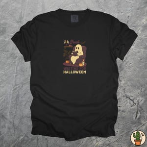 May include: Black t-shirt featuring a cartoon ghost reading a book in a chair, with the text "Just Waiting for Halloween". The ghost is in a purple chair with a candle and pumpkins.
