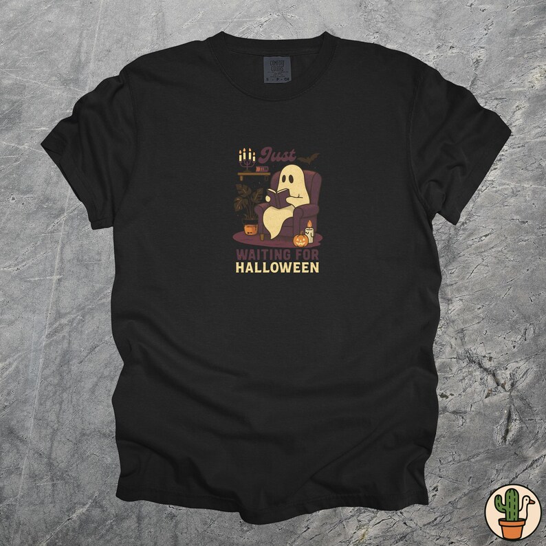 May include: Black t-shirt featuring a cartoon ghost reading a book in a chair, with the text "Just Waiting for Halloween". The ghost is in a purple chair with a candle and pumpkins.