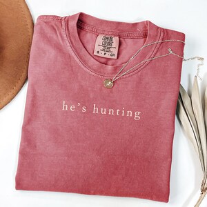 May include: A dusty rose Comfort Colors t-shirt with the phrase "he's hunting" printed in white. A silver necklace with a small pendant rests on the shirt. A brown hat and dried leaves are in the background.