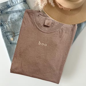 May include: A brown t-shirt with the word "boo" embroidered in white, lying on light blue denim jeans. A tan hat and a silver necklace with a pendant are also visible. The t-shirt has a crew neck and short sleeves.