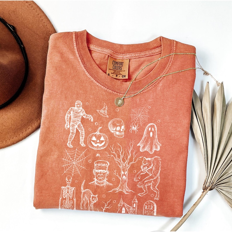 May include: An orange Comfort Colors t-shirt with a white Halloween graphic. The design includes a mummy, witch's hat, skull, spiderweb, ghost, pumpkin, Frankenstein, werewolf, cat, and tombstone. A gold necklace rests on the shirt.