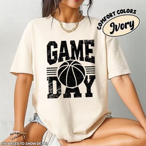 Puede incluir: Camiseta color marfil con un gr&aacute;fico negro desgastado que dice "GAME DAY" con una pelota de baloncesto en el centro. La camiseta tiene mangas cortas y cuello redondo. Las palabras "COMFORT COLORS" e "Ivory" est&aacute;n impresas arriba a la derecha.