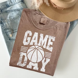 Puede incluir: Una camiseta marr&oacute;n con las palabras "GAME DAY" en letras blancas y un gr&aacute;fico de baloncesto. La camiseta est&aacute; colocada sobre unos vaqueros azul claro, un collar y un sombrero beige.