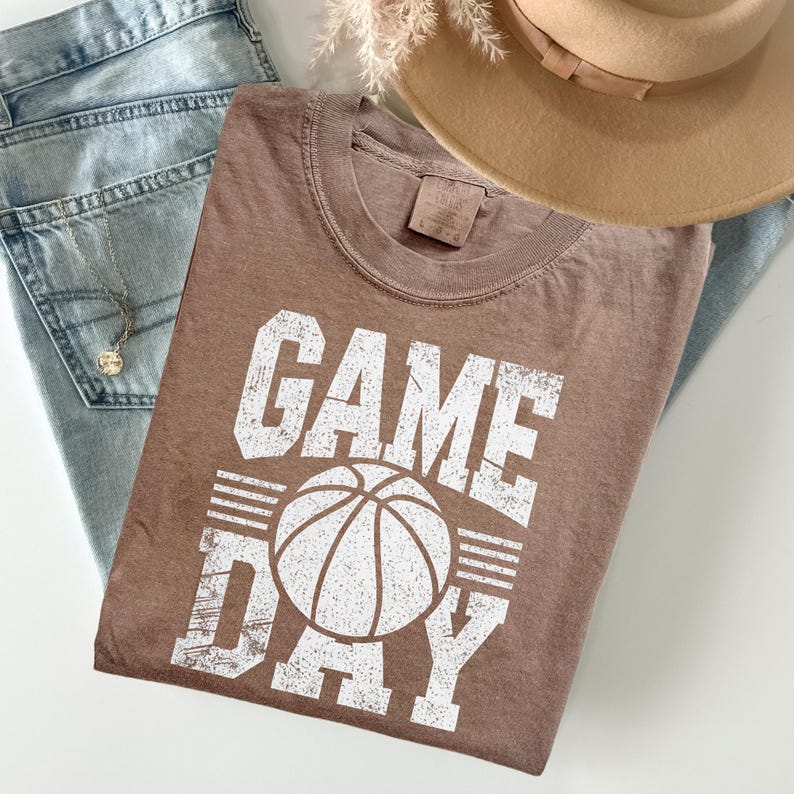 Puede incluir: Una camiseta marr&oacute;n con las palabras "GAME DAY" en letras blancas y un gr&aacute;fico de baloncesto. La camiseta est&aacute; colocada sobre unos vaqueros azul claro, un collar y un sombrero beige.