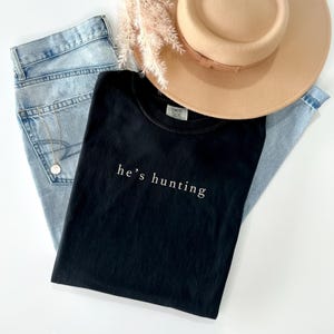 May include: A black t-shirt with the text "he's hunting" in white, paired with light-wash denim jeans, a tan fedora hat, and a delicate gold necklace. The items are arranged on a white surface with dried floral accents.