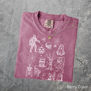 May include: Berry-coloured t-shirt with a crew neck and short sleeves. The shirt features a white graphic print of Halloween-themed illustrations, including a mummy, pumpkin, skull, and haunted house. A gold necklace rests on the shirt. The tag reads "Comfort Colors".