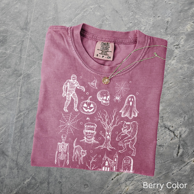 May include: Berry-coloured t-shirt with a crew neck and short sleeves. The shirt features a white graphic print of Halloween-themed illustrations, including a mummy, pumpkin, skull, and haunted house. A gold necklace rests on the shirt. The tag reads "Comfort Colors".