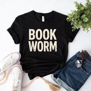 May include: Black t-shirt with the words "BOOK WORM" in a distressed, off-white font. The shirt is styled with white sneakers, denim shorts, and sunglasses. A plant and beaded necklace are also in the image.