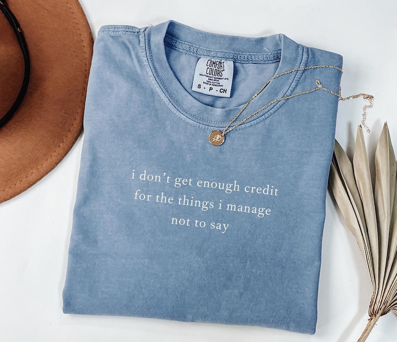 May include: A blue Comfort Colors t-shirt with the text "i don't get enough credit for the things i manage not to say" in white. A gold necklace with a pendant is draped across the shirt. A brown hat and dried plant are in the background.