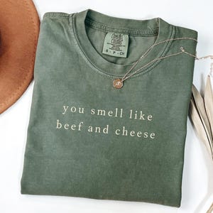 May include: An olive green t-shirt with the phrase "you smell like beef and cheese" printed in white. A silver necklace with a small pendant rests on the shirt. The shirt is folded and placed on a white surface.