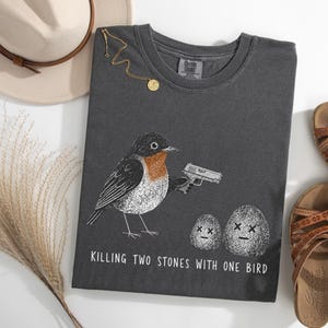 Puede incluir: Una camiseta gris carb&oacute;n con un gr&aacute;fico de un petirrojo sosteniendo una pistola y apuntando a dos piedras con ojos en forma de X. El texto debajo del gr&aacute;fico dice: "KILLING TWO STONES WITH ONE BIRD." La camiseta tiene un cuello redondo.
