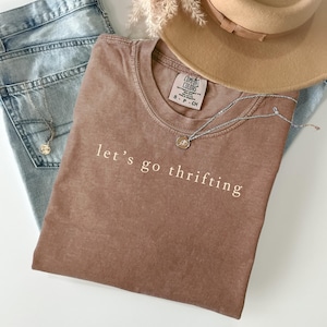 Puede incluir: Una camiseta marrón con el texto "let's go thrifting" en blanco, combinada con vaqueros azul claro, un sombrero color canela y un collar plateado. La camiseta tiene una etiqueta Comfort Colors. El collar tiene un pequeño colgante.