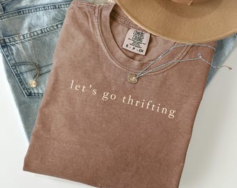 May include: A brown t-shirt with the text "let's go thrifting" in white, paired with light blue jeans, a tan hat, and a silver necklace. The shirt has a Comfort Colors tag. The necklace has a small pendant.
