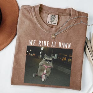 Puede incluir: Camiseta marr&oacute;n con el texto "WE RIDE AT DAWN" sobre una imagen de un mapache con gafas de sol montando un triciclo rosa. La camiseta tiene cuello redondo y mangas cortas. Un collar plateado con un peque&ntilde;o colgante est&aacute; sobre la camiseta.