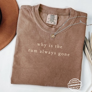 Op de afbeelding: Een bruin Comfort Colors t-shirt met de tekst "why is the rum always gone" in het wit gedrukt. Een zilveren ketting met een kleine hanger ligt op het shirt. Het shirt is opgevouwen en heeft een vintage uitstraling.