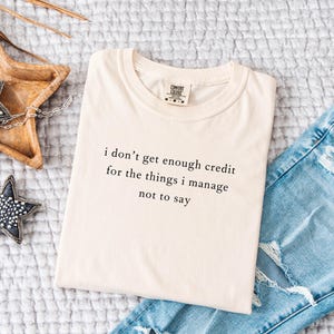 May include: Cream-colored t-shirt with the quote "i don't get enough credit for the things i manage not to say" in black text. The shirt is paired with light blue ripped jeans and decorative star-shaped ornaments.