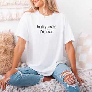May include: White t-shirt with the text "in dog years i'm dead" in a black, simple font. The shirt is a casual style, with short sleeves. The person is wearing ripped blue jeans.