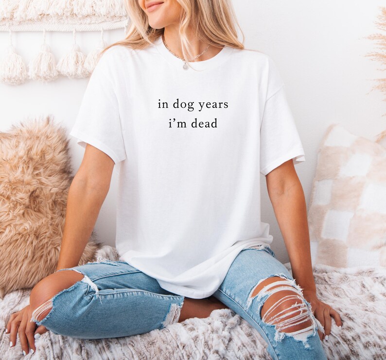 May include: White t-shirt with the text "in dog years i'm dead" in a black, simple font. The shirt is a casual style, with short sleeves. The person is wearing ripped blue jeans.