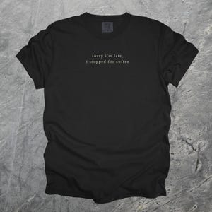 Puede incluir: Camiseta negra con la frase "sorry i'm late, i stopped for coffee" en una fuente sencilla y elegante. La camiseta est&aacute; hecha de un material suave y es adecuada para uso casual. El texto est&aacute; centrado en el pecho.