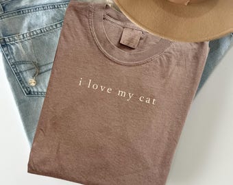 May include: A brown t-shirt with the text "i love my cat" in white lettering. The shirt is folded on top of a pair of light blue jeans, a necklace, and a tan hat.