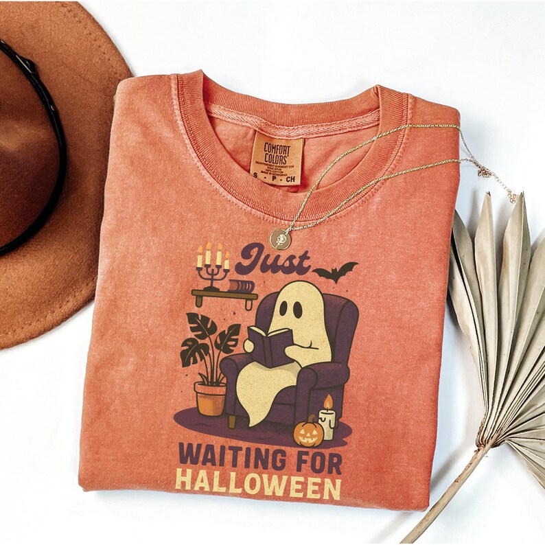 May include: Orange Comfort Colors t-shirt with a Halloween graphic. The design shows a ghost reading in a chair, with the text "Just Waiting for Halloween." A gold necklace is draped across the shirt.