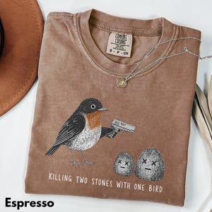 Puede incluir: Una camiseta color espresso con un gr&aacute;fico de un p&aacute;jaro sosteniendo una pistola, de pie junto a dos piedras con ojos en forma de X. El texto debajo dice "KILLING TWO STONES WITH ONE BIRD". Tiene cuello redondo y etiqueta Comfort Colors.