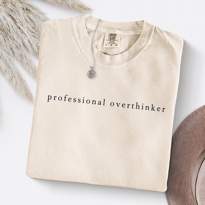 Op de afbeelding: Cr&egrave;mekleurig Comfort Colors T-shirt met de zwarte tekst "professional overthinker". Een zilveren ketting met een ronde hanger ligt op het shirt. Het shirt is netjes op een wit oppervlak gevouwen.