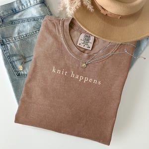 May include: A brown t-shirt with the words "knit happens" printed in white. The shirt is paired with light blue jeans, a tan hat, and a silver necklace with a pendant. The shirt has a Comfort Colors tag.