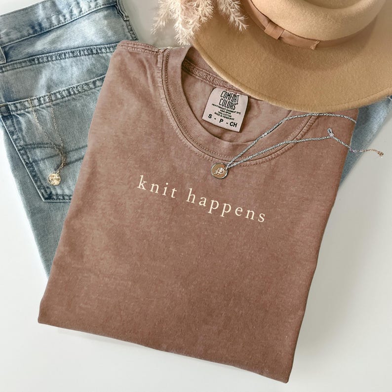 May include: A brown t-shirt with the words "knit happens" printed in white. The shirt is paired with light blue jeans, a tan hat, and a silver necklace with a pendant. The shirt has a Comfort Colors tag.