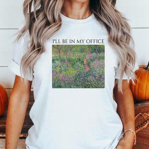 May include: White t-shirt with the text "I'll be in my office" printed on the front. The text is centered and in black. The shirt features a painting of a garden with purple flowers and green trees.