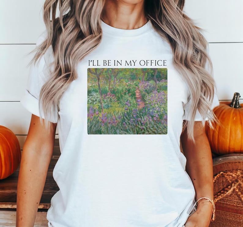 May include: White t-shirt with the text "I'll be in my office" printed on the front. The text is centered and in black. The shirt features a painting of a garden with purple flowers and green trees.
