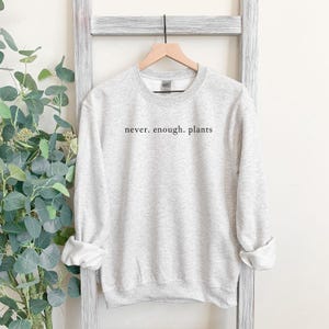 May include: A light gray sweatshirt with the text "never. enough. plants" in black. The sweatshirt is hanging on a wooden hanger against a white background. The sleeves are rolled up. The overall theme is plant-related.