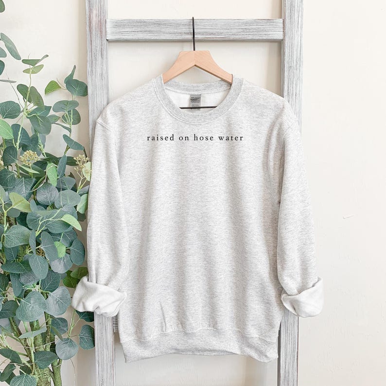 May include: A light gray sweatshirt with the phrase "raised on hose water" printed in black. The sweatshirt is hanging on a wooden hanger against a white background.