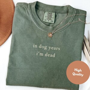 May include: An olive green t-shirt with the text "in dog years I'm dead" in white. A gold necklace with a pendant is on the shirt. A brown hat is visible in the upper left corner. A "High Quality" sticker is on the bottom right.
