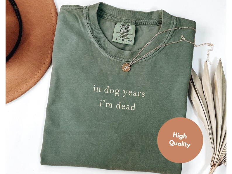 May include: An olive green t-shirt with the text "in dog years I'm dead" in white. A gold necklace with a pendant is on the shirt. A brown hat is visible in the upper left corner. A "High Quality" sticker is on the bottom right.