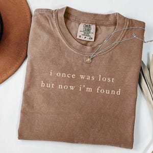 May include: A brown Comfort Colors t-shirt with the text "i once was lost but now i'm found" in white. A silver necklace with a small pendant rests on the shirt. The shirt is folded and has a tag.