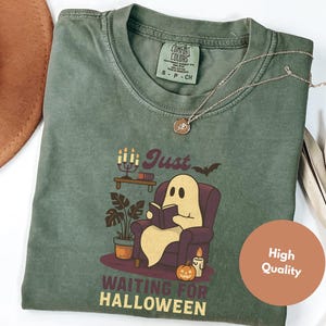 May include: Green t-shirt featuring a ghost reading a book, with the text "Just Waiting for Halloween." The design includes a bat, candles, and a pumpkin. A silver necklace is draped on the shirt. The t-shirt has a Comfort Colors label.
