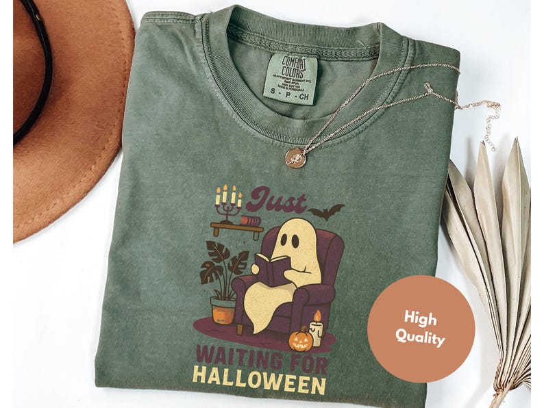May include: Green t-shirt featuring a ghost reading a book, with the text "Just Waiting for Halloween." The design includes a bat, candles, and a pumpkin. A silver necklace is draped on the shirt. The t-shirt has a Comfort Colors label.