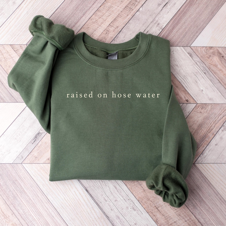 May include: An olive green sweatshirt with the phrase "raised on hose water" printed in white. The sweatshirt has a crew neck and long sleeves with cuffed wrists. The garment is folded on a light brown wooden surface.