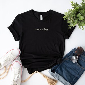 May include: Black t-shirt with the text "mom vibes" in white lettering. The shirt is laid flat on a white surface with a pair of white sneakers, a wooden bead necklace, and a pair of denim shorts.