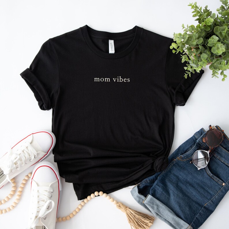 May include: Black t-shirt with the text "mom vibes" in white lettering. The shirt is laid flat on a white surface with a pair of white sneakers, a wooden bead necklace, and a pair of denim shorts.