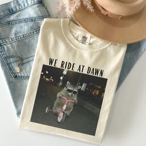 Puede incluir: Camiseta color crema con el texto "WE RIDE AT DAWN" sobre un gr&aacute;fico de un mapache con gafas de sol montando un triciclo rosa. La camiseta est&aacute; colocada sobre unos vaqueros azules y un sombrero beige.