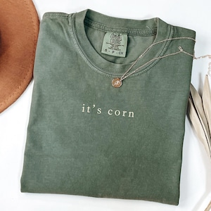 Op de afbeelding: Een olijfgroen t-shirt met de witte tekst "it's corn". Het shirt is opgevouwen en heeft een Comfort Colors-label bij de halslijn. Een gouden ketting met een hanger ligt op het shirt. Een bruine hoed is gedeeltelijk zichtbaar.