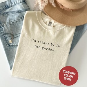 May include: Cream-colored t-shirt with the text "i'd rather be in the garden" in black. The shirt is paired with light blue jeans, a tan hat, and a silver necklace. A red circle with white text reads "Comfort Color Shirt".
