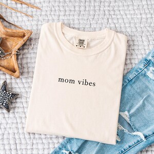 May include: A cream colored t-shirt with the text "mom vibes" printed in black on the front. The t-shirt is laying on a white textured surface with a pair of blue jeans and a wooden star-shaped tray with two decorative stars.