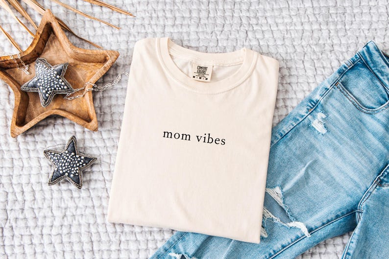 May include: A cream colored t-shirt with the text "mom vibes" printed in black on the front. The t-shirt is laying on a white textured surface with a pair of blue jeans and a wooden star-shaped tray with two decorative stars.